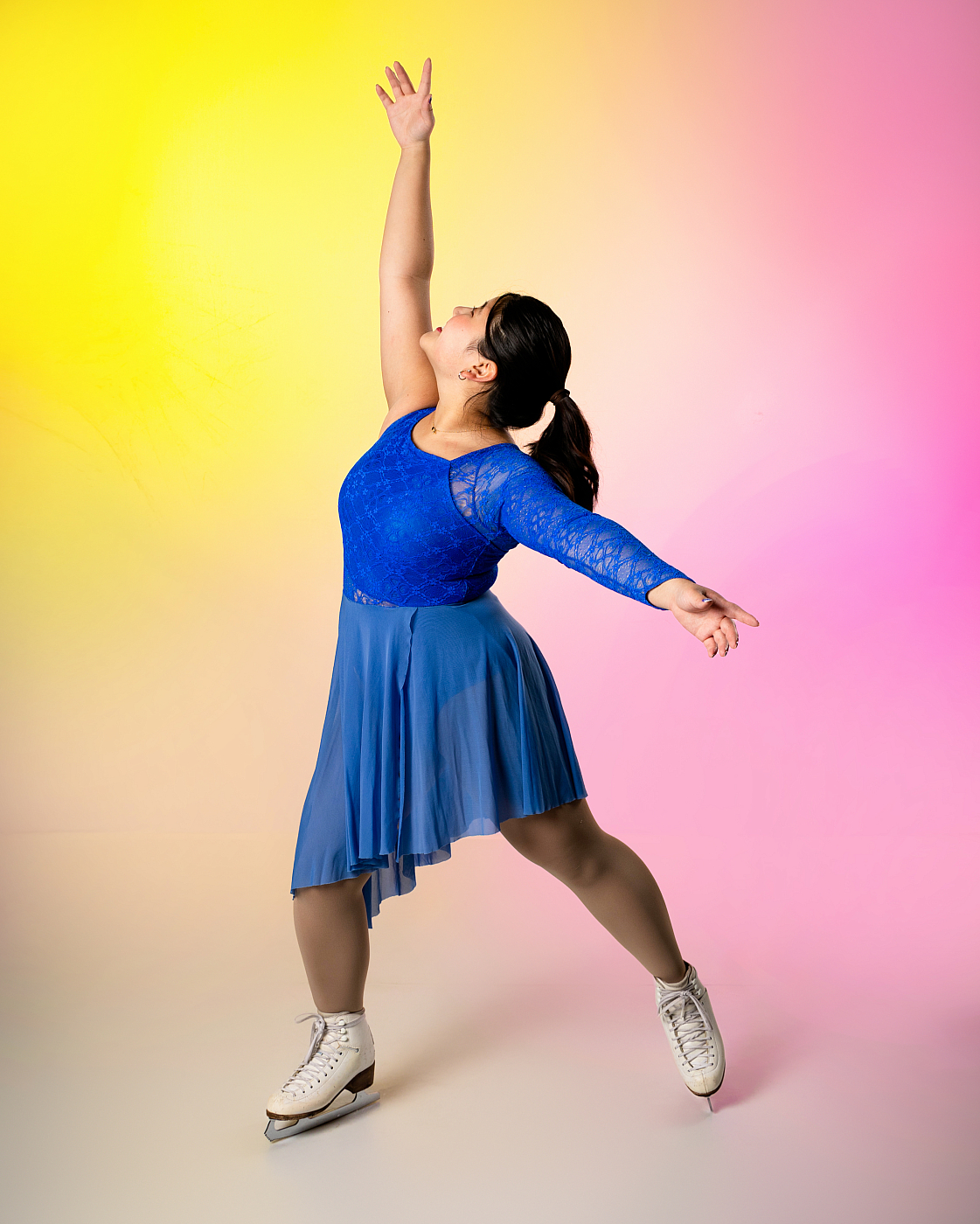 Teenage girl posing in blue leotard with skirt, wearing figure skates for indoor portrait with pink and yellow background.