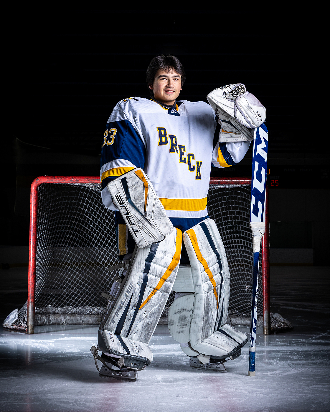 Teenage boy in blue, yellow, and white hockey uniform posing in front of hockey goal for sports portrait.