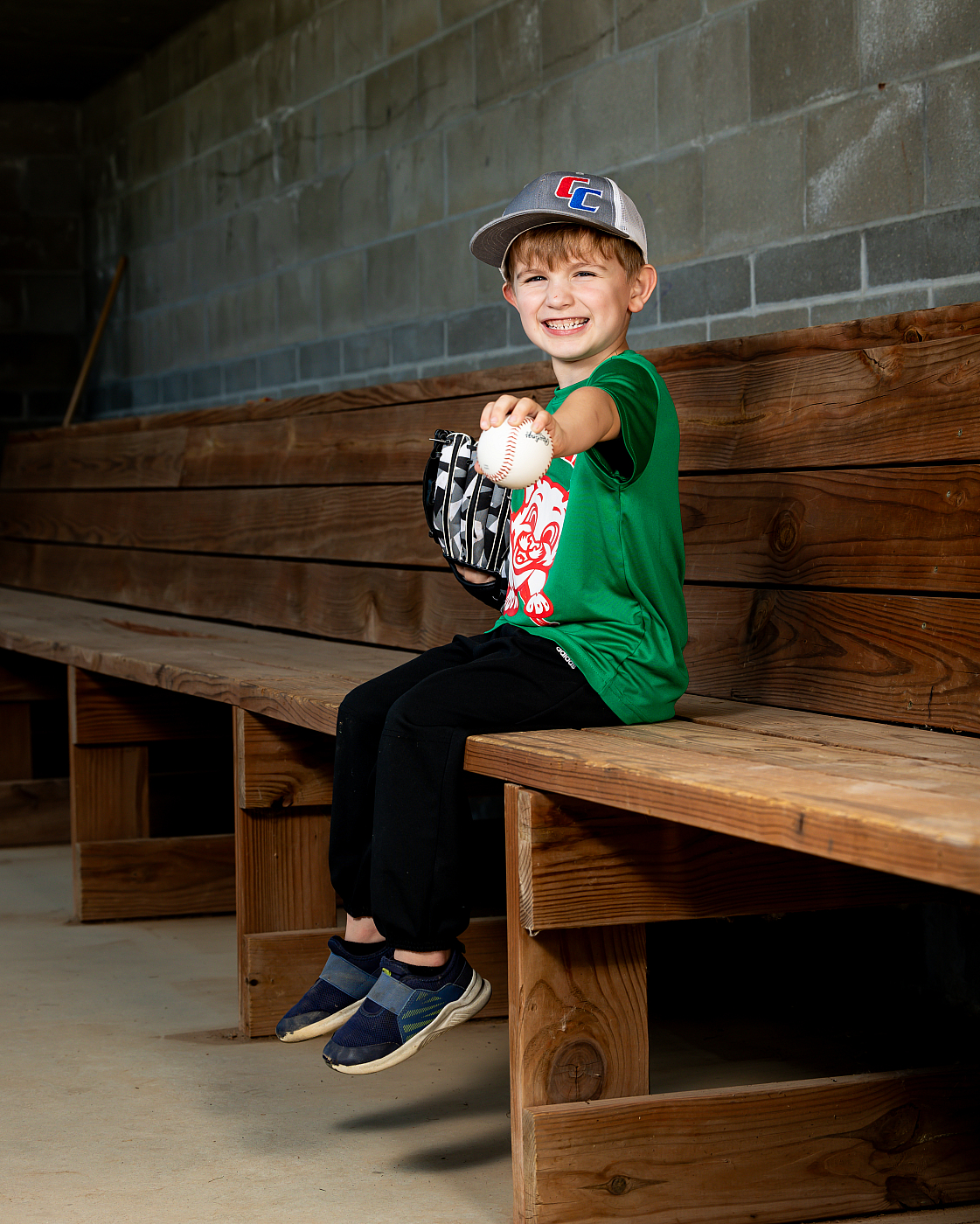 Little boy in green shirt, sitting on bench, holding out baseball for T-ball sports portrait.