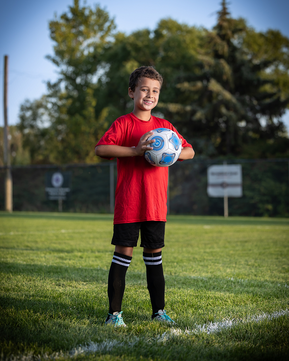 Little boy holding soccer ball on soccer field for sports portrait, wearing red soccer uniform.
