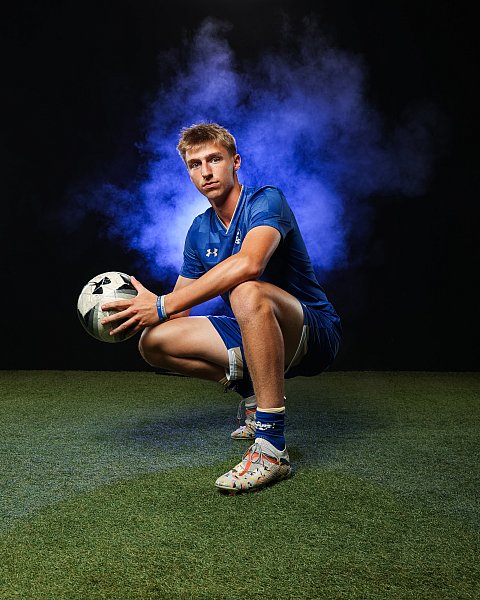 male soccer player in blue uniform squatting and holding soccer ball, blue smoke in background
