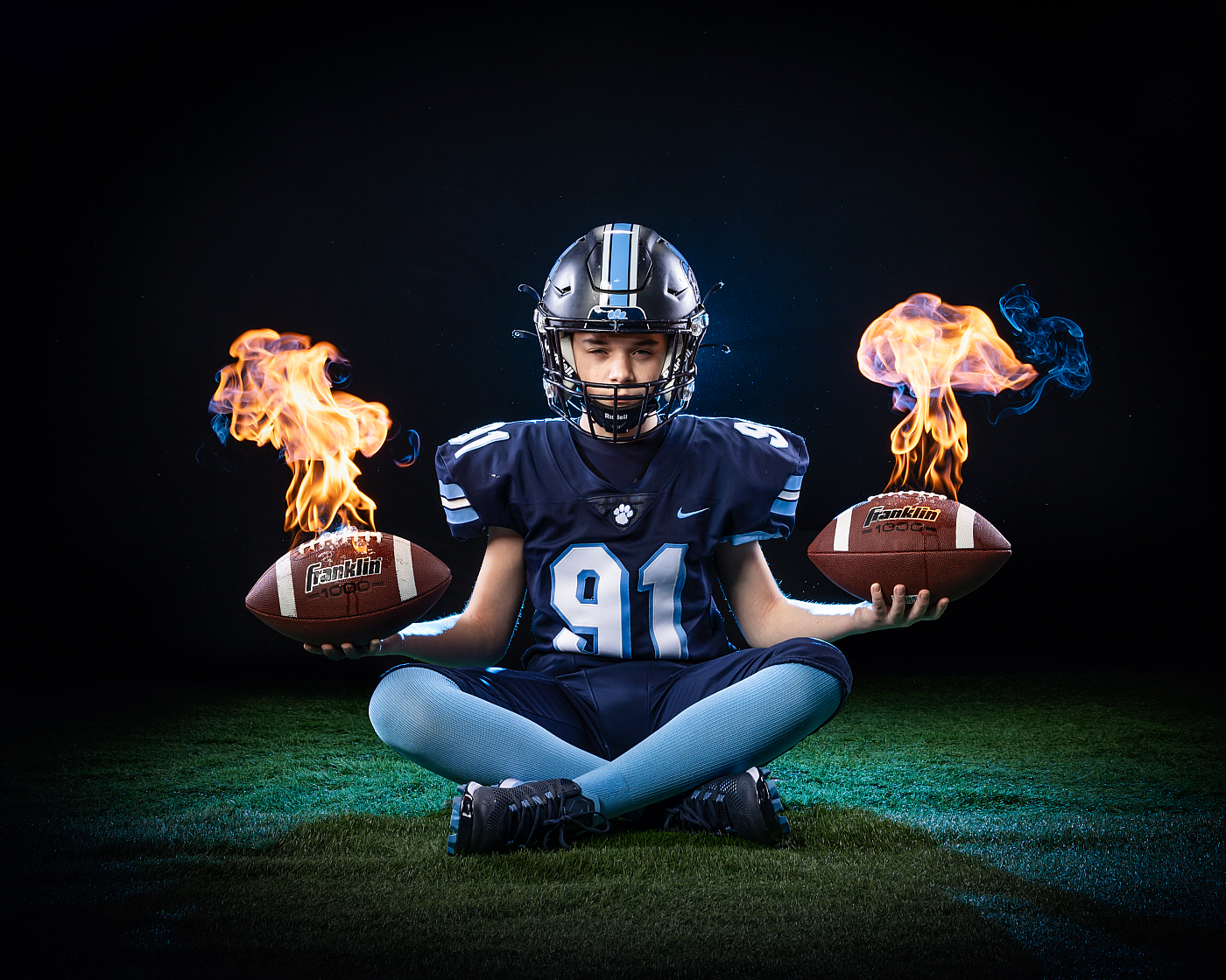boy in blue football uniform sitting holding two footballs in flames