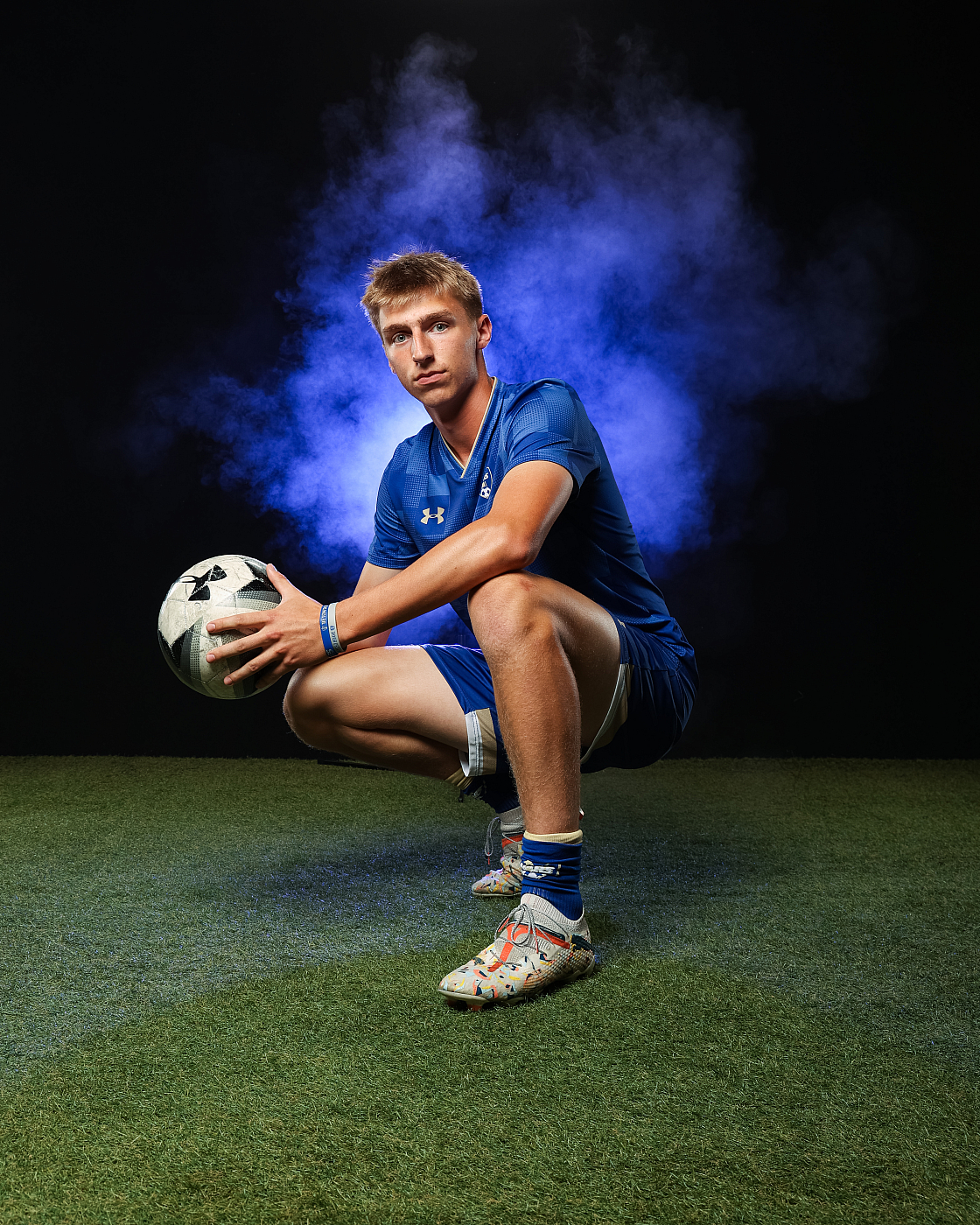 male soccer player in blue uniform squatting and holding soccer ball, blue smoke in background